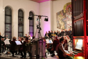 Der Musikverein im Altarraum der Rettigheimer Kirche beim Winterkonzert. Im Vordergrund Tim Brucker an der Kirchenorgel. Im der Mitte das Blasorchester. Im Hintergrund die Sänger*innen von "New Voice"